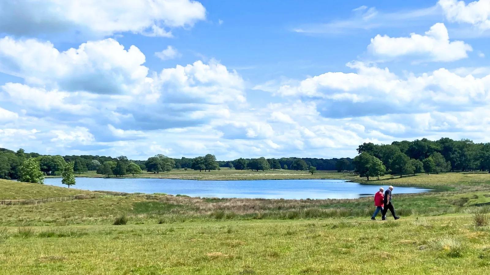 Two walkers crossing grassland beside Tatton Mere under a bright sky