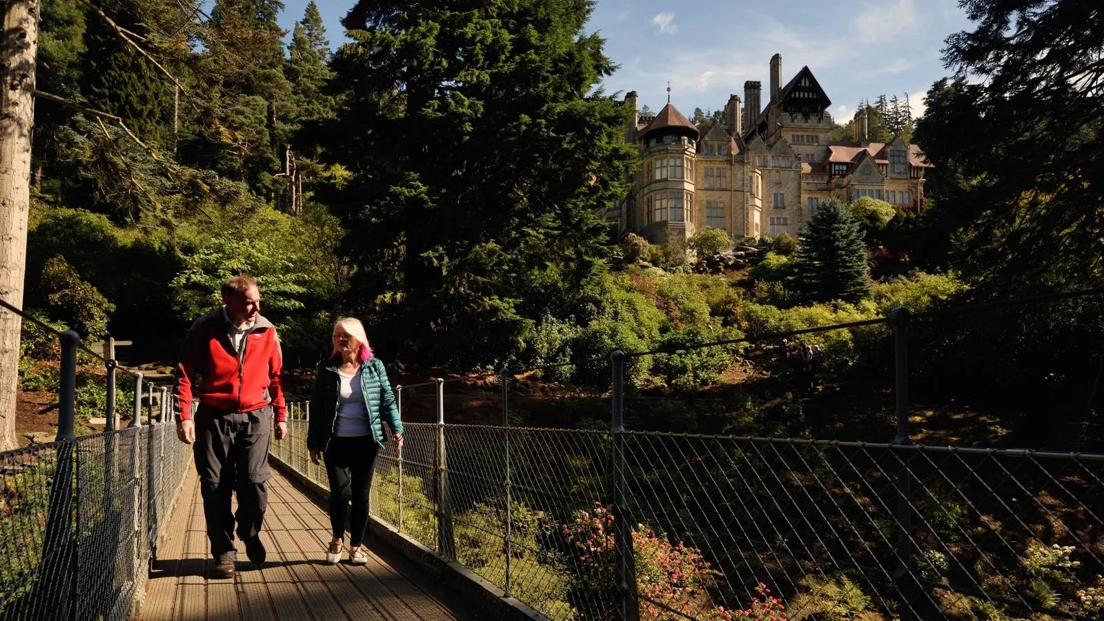 Two visitors crossing the Iron Bridge below Cragside House and its wooded rock garden