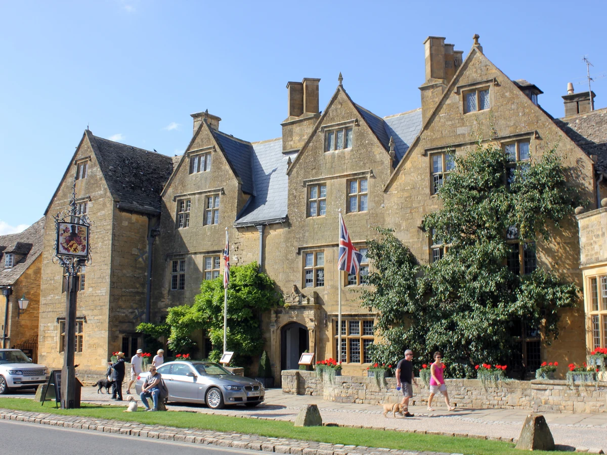 The sunlit stone frontage of The Lygon Arms with people walking dogs in Broadway