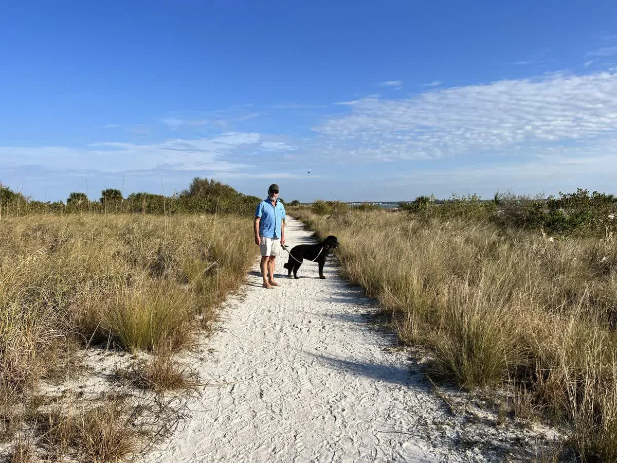 A visitor walks a leashed dog along a sandy trail toward the shore at Honeymoon Island State Park.