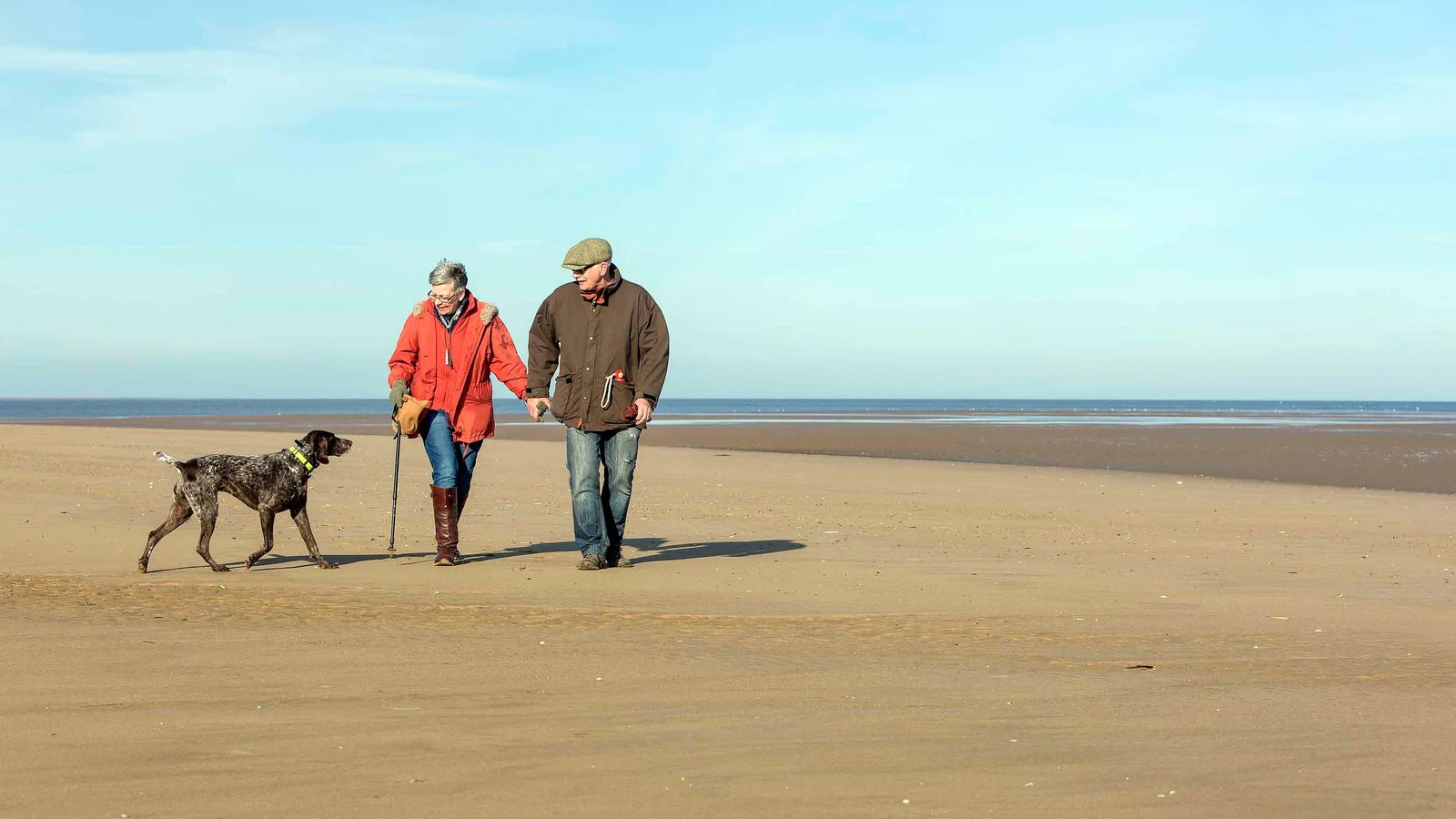 Two people walking a dog across Brancaster Beach