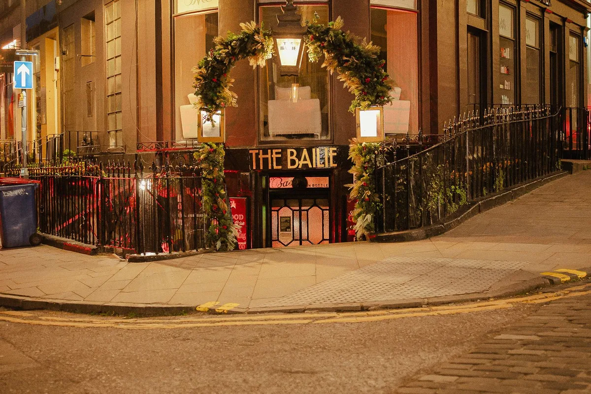 Illuminated corner entrance of The Bailie, with a low doorway and pavement railings.